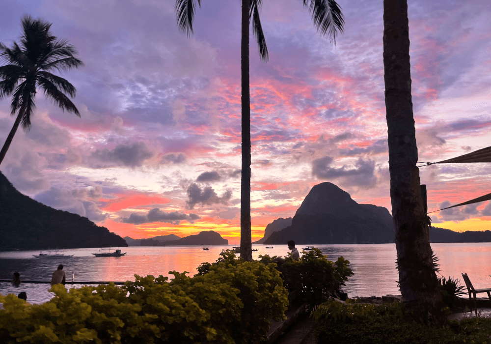 Sunset over El Nido framed by palm trees and anchored boats.