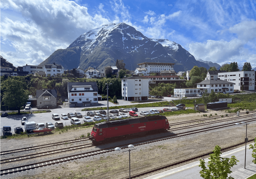 A red train crossing a mountain town below a snow-tipped peak.